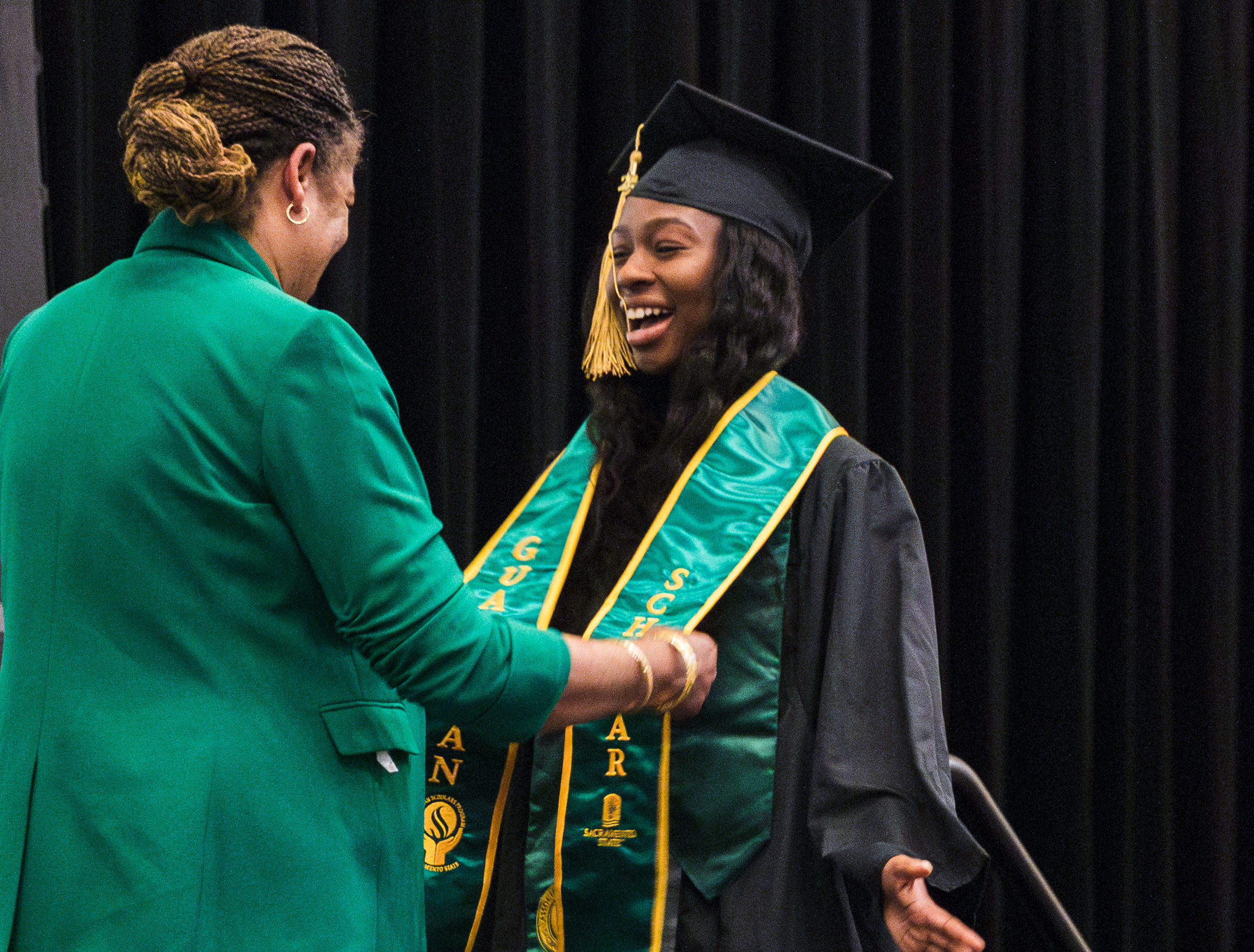 A woman in green blazer drapes a stole around a woman in graduation cap and gown.